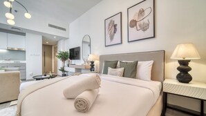 A modern hotel room with a neatly made bed, a sitting area with a chair, a small table, and a lamp. There is a kitchen area visible in the background. - Classic Studio at Mesk 1 Midtown Dubai Production City by Deluxe Holiday Homes (Dubai)