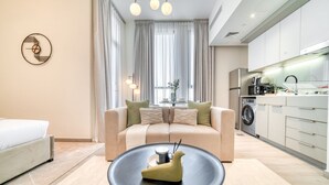 A modern hotel room with a sofa, a dining table with chairs, a kitchen area with appliances, and a large window with curtains. - Classic Studio at Mesk 1 Midtown Dubai Production City by Deluxe Holiday Homes (Dubai)