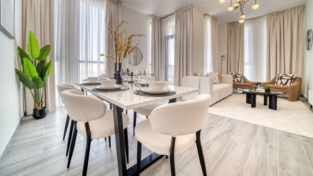 A modern dining area with a white table set for four, surrounded by beige chairs. A large potted plant is on the left, and a dining table lamp is on the right.