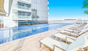A rooftop pool area with lounge chairs and a modern building in the background. - Delightful Studio at Carson A DAMAC Hills Dubailand by Deluxe Holiday Homes (Dubai)