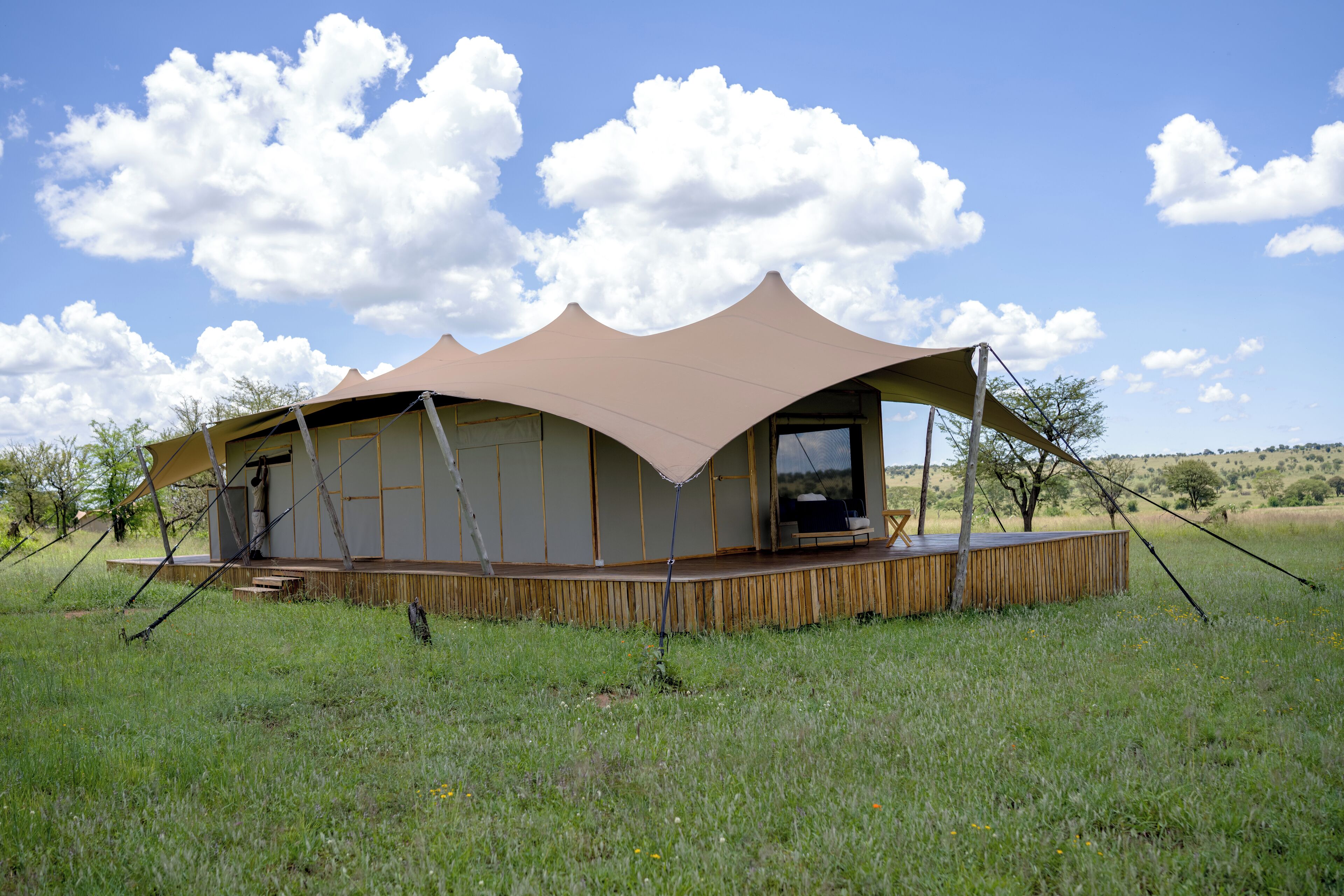 A large, open-air structure with a flat roof and a wooden deck, surrounded by a grassy field and trees.