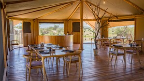 A spacious tented dining area with wooden tables and chairs, a central bar, and large windows offering views of the outdoors.