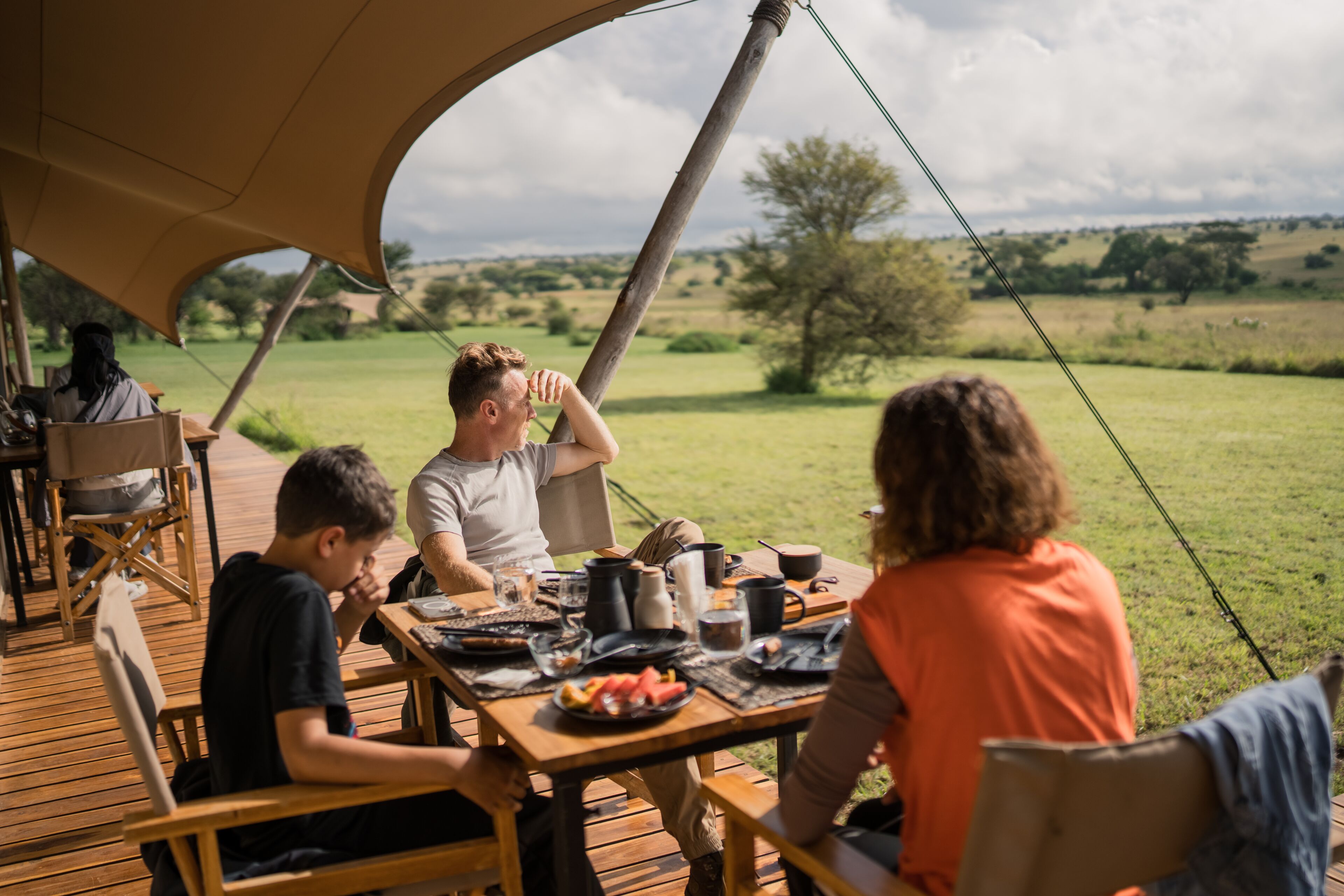 A group of people dining at an outdoor wooden table with a view of a grassy field and trees.