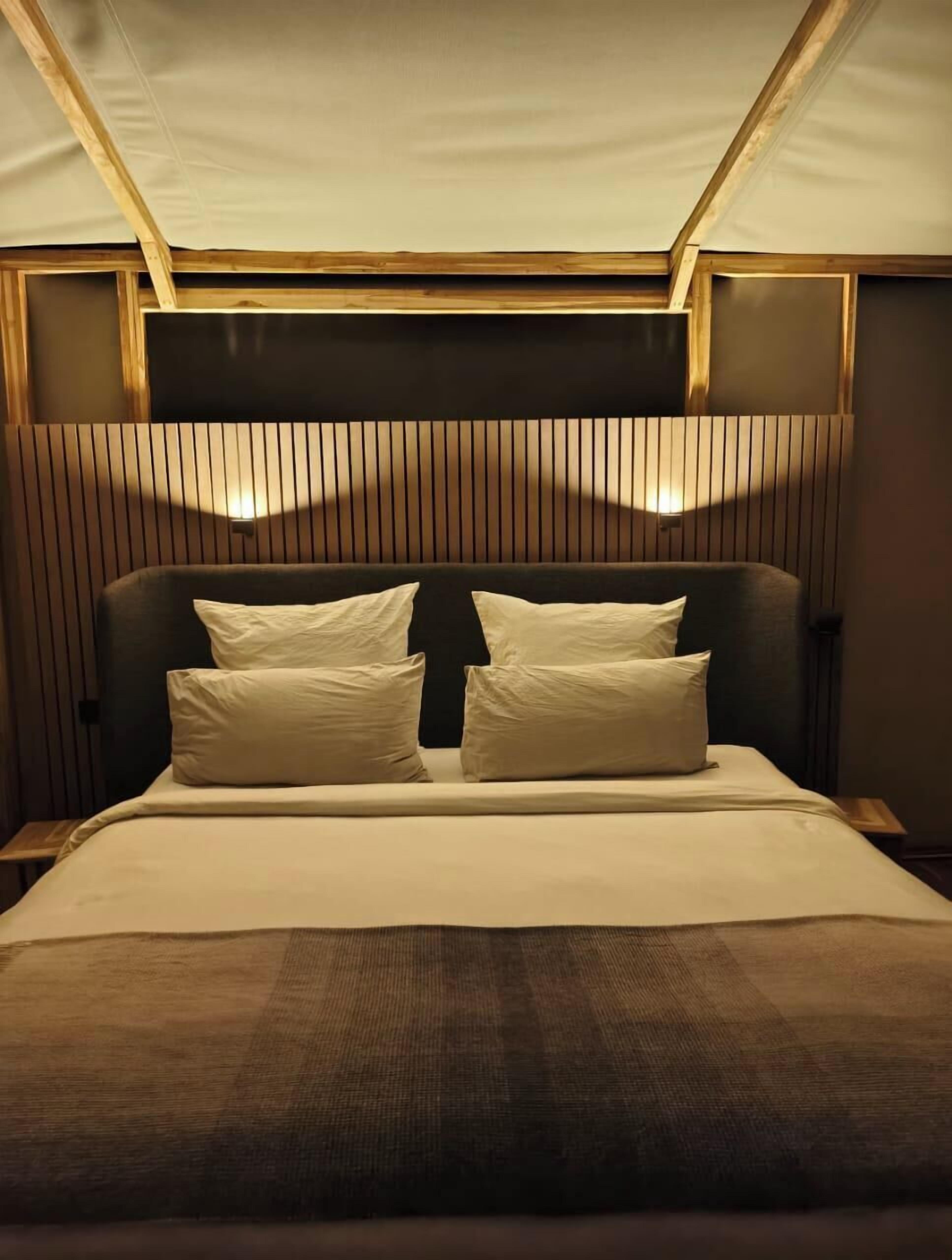 A hotel room with a bed, two bedside tables, a headboard with vertical slats, and a ceiling with a wooden beam structure.
