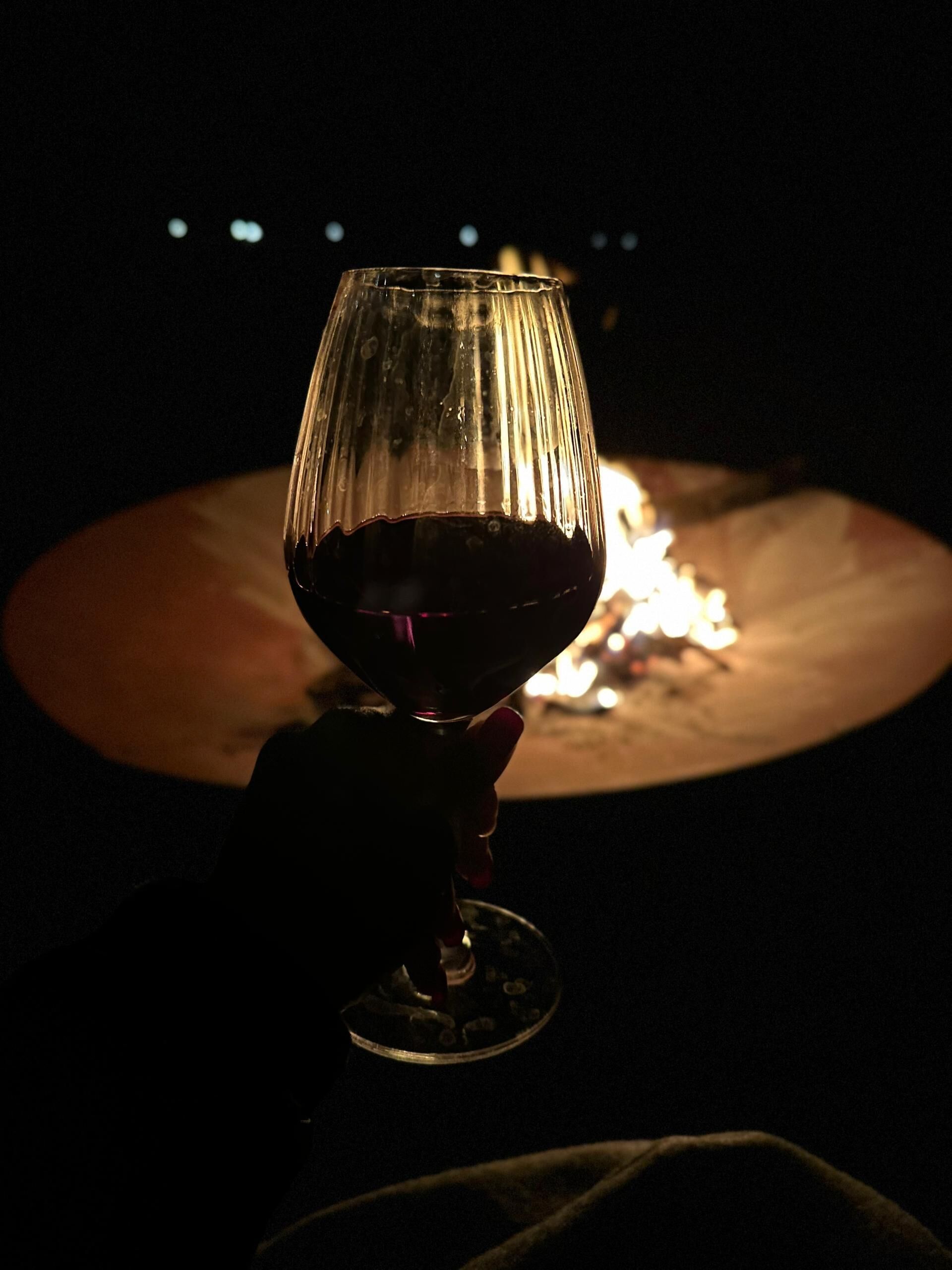 A glass of red wine held in a dimly lit setting with a fire in the background.