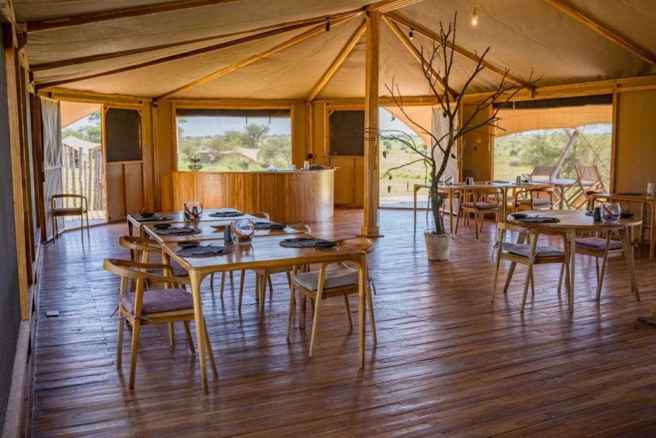 A tented dining area with wooden tables and chairs, a central bar, and a view of the outdoors.