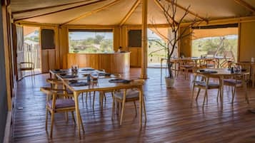 A tented dining area with wooden tables and chairs, a central bar, and a view of the outdoors.
