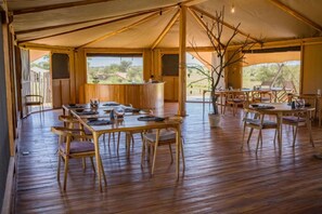 Un espace de restauration aménagé sous une tente, avec des tables et des chaises en bois, un bar central et une vue sur l’extérieur.