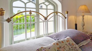 A bedroom with a large window, a bed with floral bedding, and a bedside table with a lamp. - Chichester Lodge -  a cottage that sleeps 8 guests  in 4 bedrooms (Chichester)
