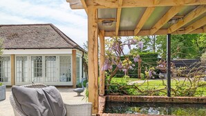 A covered patio with wicker furniture, a brick wall, and a view of a house with large windows and a garden. - Chichester Lodge -  a cottage that sleeps 8 guests  in 4 bedrooms (Chichester)