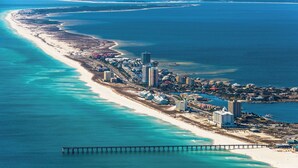 Vista aérea de una ciudad costera con una extensa playa de arena, un muelle que se adentra en el agua y varios edificios altos a lo largo de la costa.