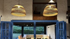 Zona pranzo con tavolo in legno, lampade a sospensione in vimini e vista su un giardino attraverso porte blu aperte.