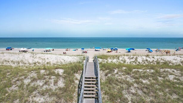 Une plage avec une promenade en bois menant au rivage, des dunes de sable et des parasols de plage.