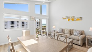 A modern living room with a wooden dining table, beige sofa, and a view of the ocean through large windows.