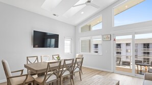 A modern dining area with a wooden table and chairs, a ceiling fan, and large windows offering a view of the outside.