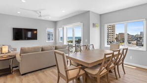 A modern living room with a sofa, dining table, and chairs. There is a TV mounted on the wall, and large windows offering a view of the city. - Seadragon 2 | Pet-Friendly w/ Rooftop Deck + Ocean Views! (Carolina Beach)