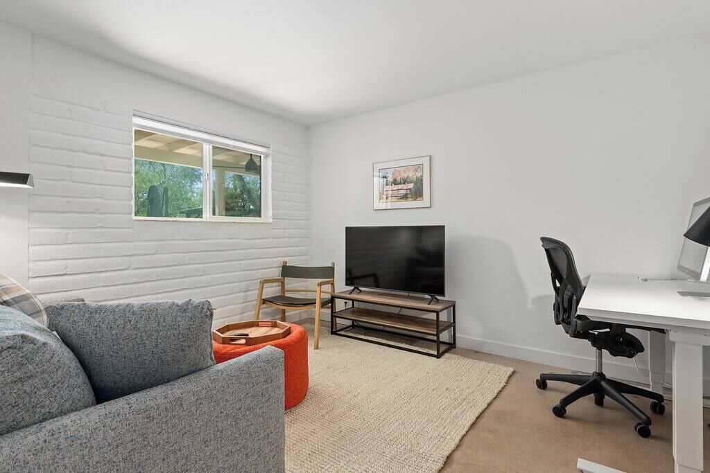 A modern living room with a grey sofa, a wooden coffee table, a TV on a wooden stand, a desk with a computer, and a window with a view of greenery.