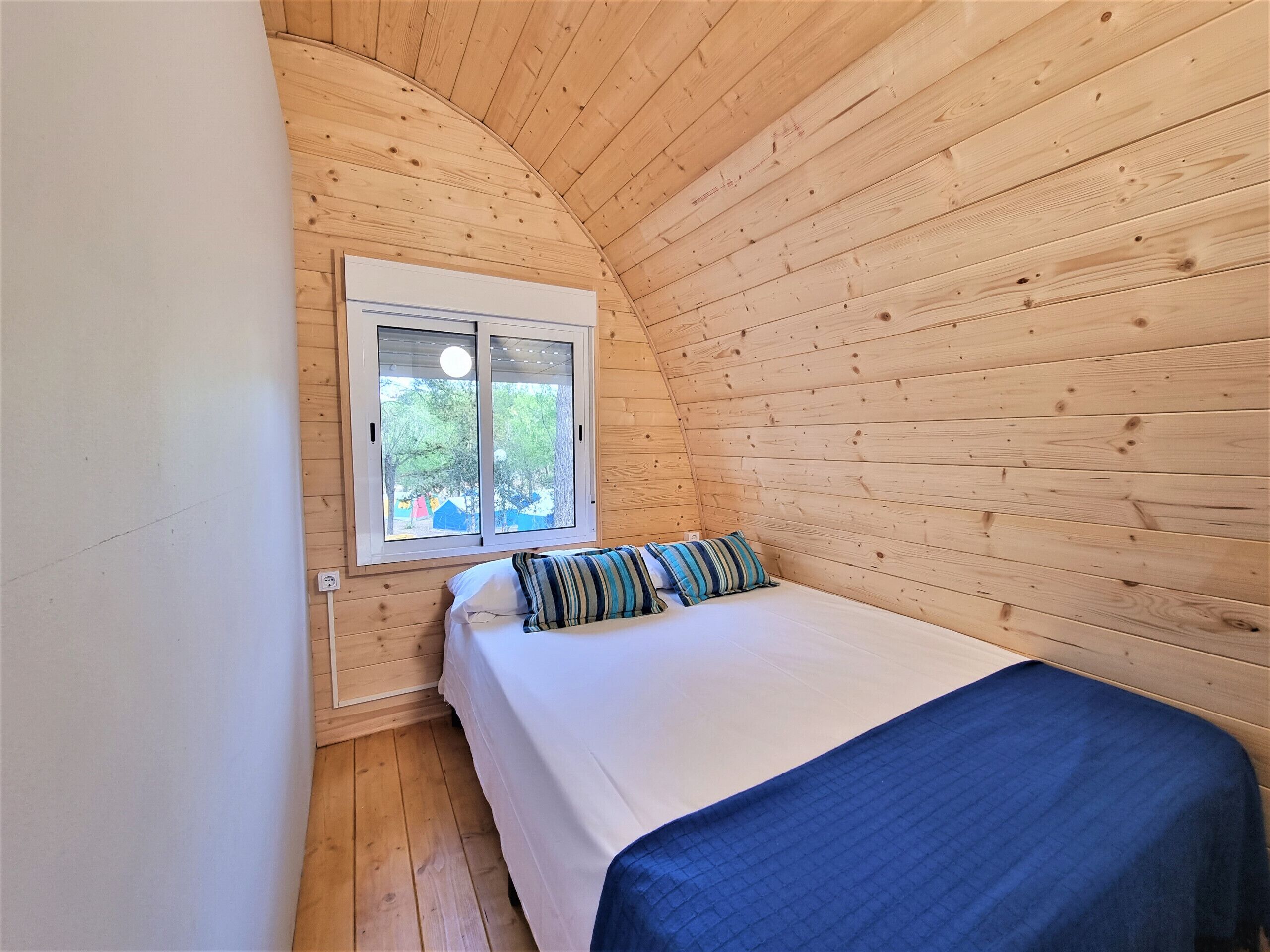 Een kleine, gezellige kamer met houten wanden, een enkel bed met een blauwe deken, en een raam met uitzicht op bomen.