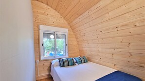 Une petite pièce chaleureuse aux murs en bois, avec un lit simple recouvert d’une couverture bleue et une fenêtre donnant sur les arbres.