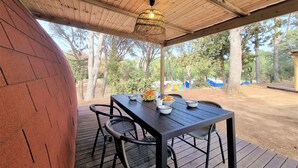 A covered outdoor dining area with a table and chairs, a hanging light fixture, and a view of trees and tents in the background. - Nomad Paraiso: Beautiful cozy and familiar bungalow (L'Escala)