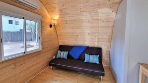 A wooden cabin room with a sofa, a wall-mounted lamp, and a window overlooking a fenced yard. - Nomad Paraiso: Beautiful cozy and familiar bungalow (L'Escala)