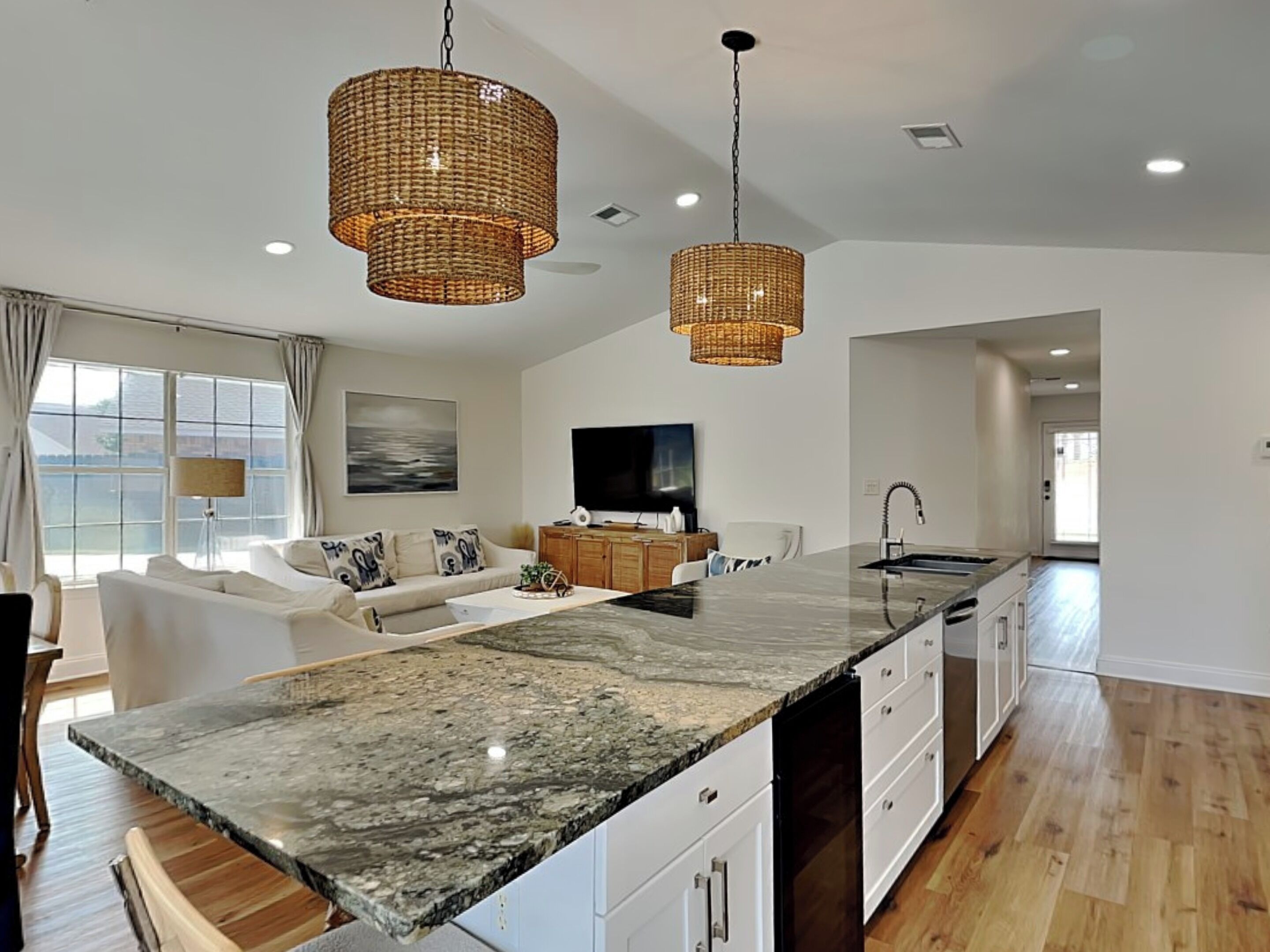 A modern kitchen with a large island, two hanging wicker chandeliers, a flat-screen TV, and a view of the city through large windows.