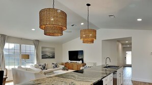 A modern kitchen with a large island, two hanging wicker chandeliers, a flat-screen TV, and a view of the city through large windows. - Seaspray | Private Pool + 4 Bikes (Miramar Beach)