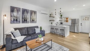 Ein modernes Wohnzimmer mit grauem Sofa, einem Holztisch und einer Küchenzeile mit weißer Kochinsel und Barhocker.