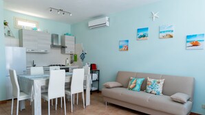 A modern kitchen with white cabinets, a dining table with chairs, a beige sofa, and three beach-themed wall art pieces. - Stella Marina apartment, vacation home in Trapani, a few steps from the sea  (Trapani)