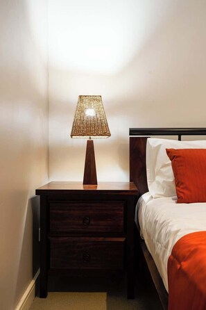Un lit bien fait, avec une literie blanche et orange, une table de chevet en bois avec une lampe, et une tête de lit en bois.