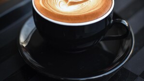 A cup of coffee with latte art on a black saucer on a reflective surface. - Roomshala 059 Sp Lodge (New Delhi)