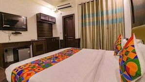 A hotel room with a bed, a TV mounted on the wall, a desk with a chair, and a window with curtains. - Roomshala 059 Sp Lodge (New Delhi)