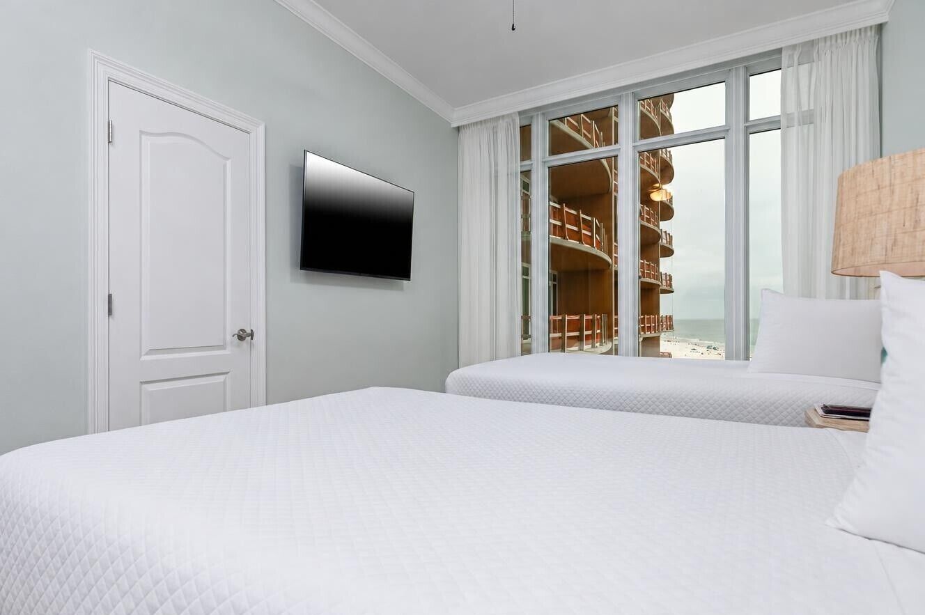 A hotel room with a bed, a TV, a window with a view of a beach, and a bookshelf.