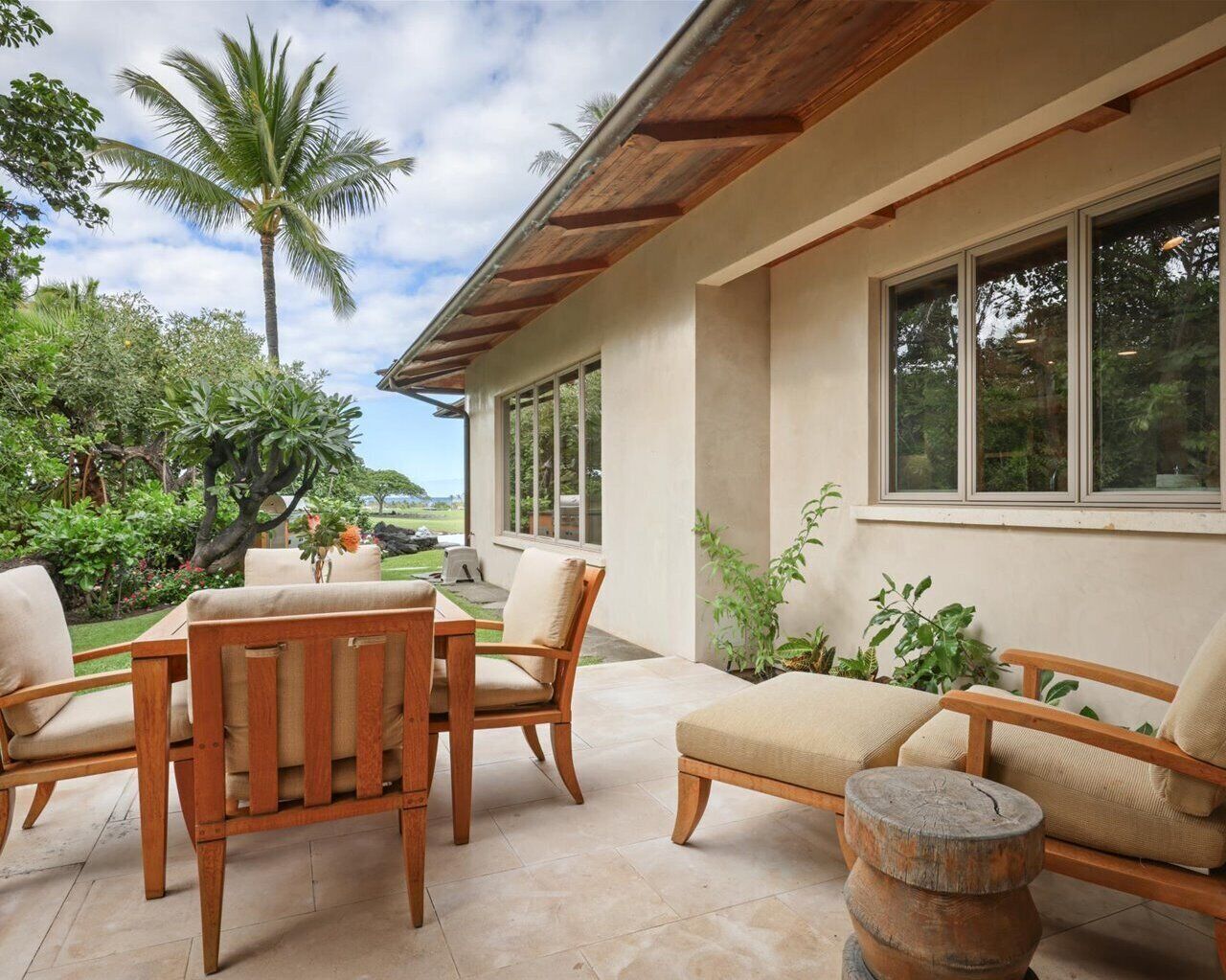 An outdoor patio with wooden furniture, a stone table, and a view of a lush garden and palm tree.