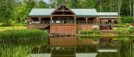 A rustic lodge with a green roof, wooden exterior, and a deck overlooking a pond.