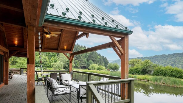Una terraza de madera con mobiliario exterior, un espacio cubierto con techo metálico y vista a un lago y un bosque.