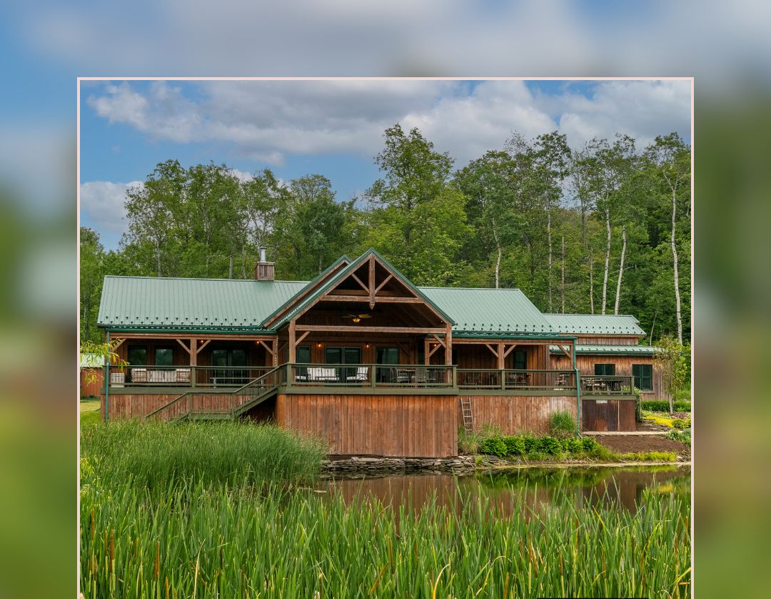 Une cabane rustique avec un toit végétalisé, un revêtement extérieur en bois et une jetée qui s’avance sur un étang, le tout entouré d’herbes hautes et d’arbres.