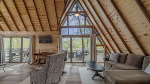 Un loft amplio con techo alto, vigas de madera, un gran ventanal corredero que da a una terraza exterior, un sofá, un sillón estampado y una mesita.