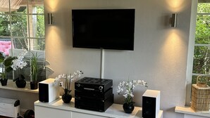 A modern living room with a flat-screen TV mounted on the wall, a white entertainment unit with electronic devices, and a large window offering a view of greenery outside. - ERMELO Hideaway  (Ermelo)