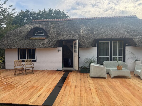 Un bâtiment doté d’un toit de chaume, de murs blancs et d’une terrasse en bois aménagée avec des meubles en osier.