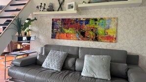 A modern living room with a grey leather sofa, a colorful abstract painting, a staircase, and a small shelf with decorative items. - ERMELO Hideaway  (Ermelo)