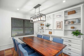 A dining area with a wooden table, blue chairs, white shelving with decorative items, and a hanging light fixture.