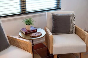Un rincón acogedor con dos sillones de mimbre, una mesita redonda con una planta y un libro titulado “Texas Whiskey”, y una ventana con persianas.