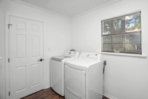 A laundry room with two washing machines, a dryer, a white door, and a window with a view of trees. - A Pecan Retreat-Fire pit- 3 Blocks to Main St! (Fredericksburg)