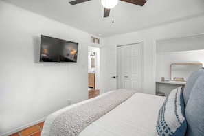 A bedroom with a bed, a TV mounted on the wall, a ceiling fan, and a door leading to another room.