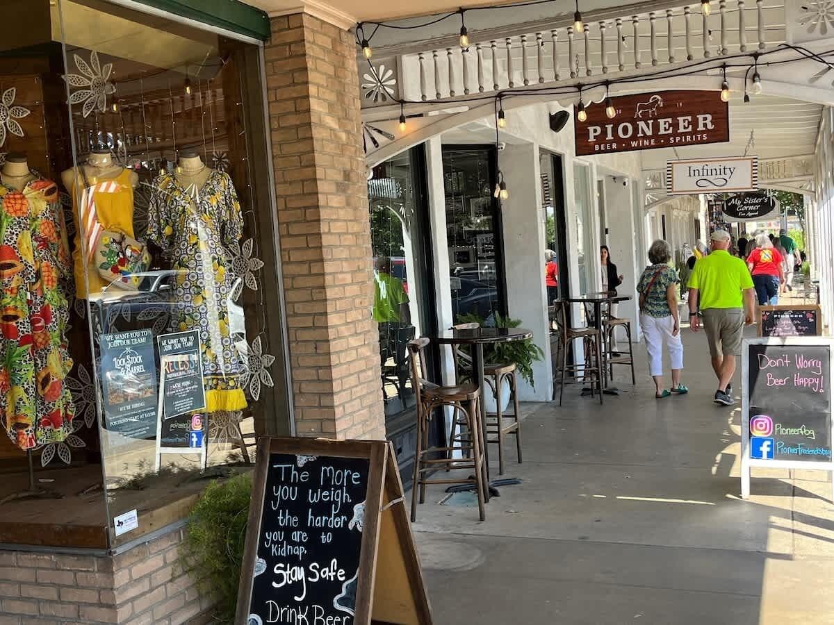 A storefront with mannequins displaying colorful clothing, a sign for 'Pioneer Beer, Wine, Spirits', and a chalkboard with a safety message.