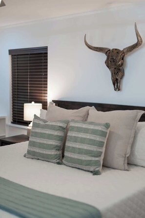 Un dormitorio con una cama grande, almohadones a rayas, un cabecero, un cráneo de toro montado en la pared, una mesita de noche con una lámpara y una ventana con persianas.