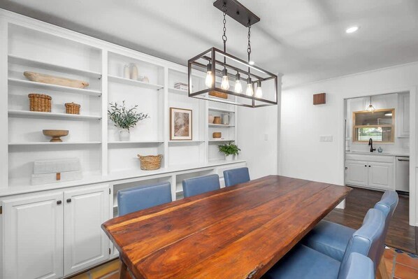 Un comedor moderno con una mesa de madera y sillas azules, estanterías blancas con objetos decorativos y una lámpara colgante.