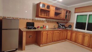 A kitchen with wooden cabinets, a refrigerator, a microwave, and a window with green blinds. - Appartement Spacieuse et Propre à Louer (Martil)
