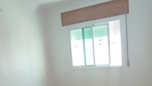 A room with a large window, white walls, and a brown tiled floor. - Appartement Spacieuse et Propre à Louer (Martil)